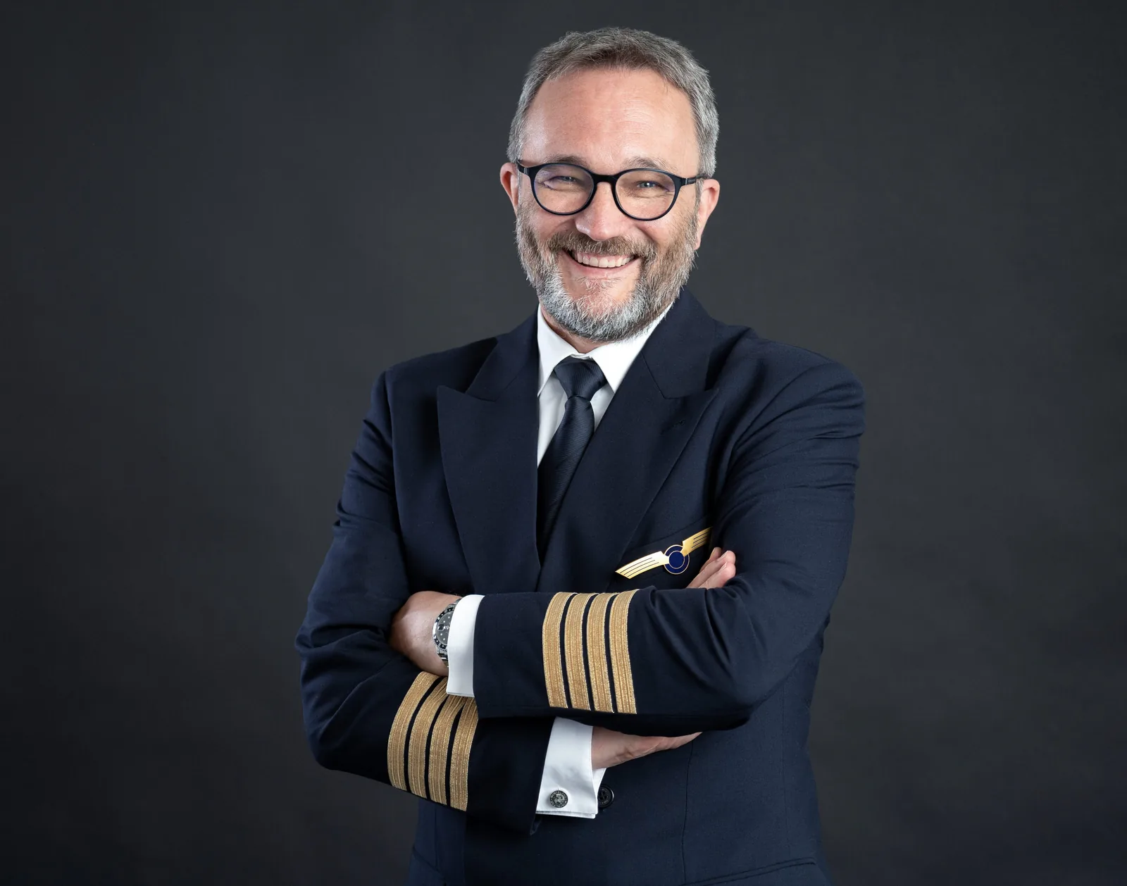 Flugkapitän in dunkler Uniform mit vier Streifen und Schwingenabzeichen, Arme verschränkt, vor anthrazitfarbenem Hintergrund