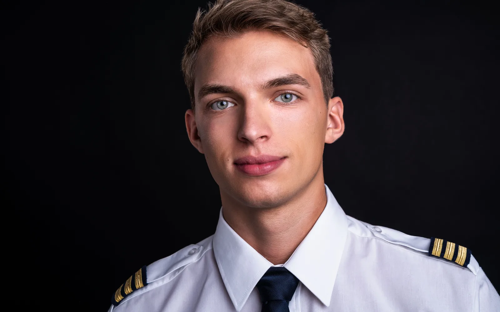 Junger Pilot in weißem Uniformhemd mit Krawatte und Schulterklappen, ruhiger Blick, vor schwarzem Hintergrund