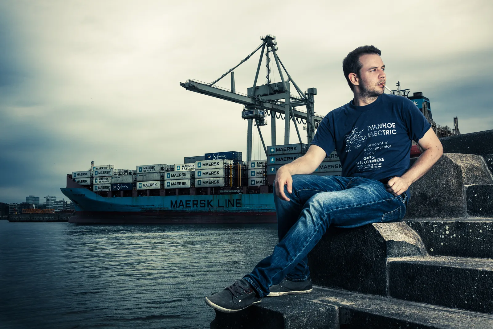 Mann sitzt auf einer Kaimauer im Hamburger Hafen, dahinter ein Containerschiff der Maersk-Linie und die Containerbrücken