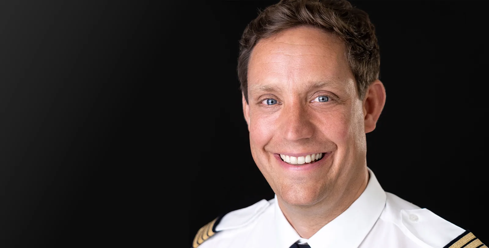 Pilot in weißem Uniformhemd mit Schulterklappen, lachend vor schwarzem Hintergrund